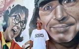 Se observa al artista urbano posando para la foto y detrás de él el mural donde se aprecia a el Chavo y la Chilindrina